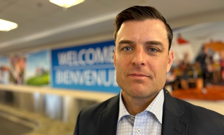 A man with short hair and a suit stands inside an airport terminal looking at the camera.