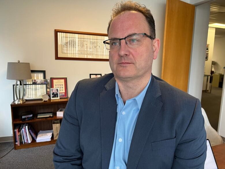 A man with brown hair and glasses wearing a blue shirt and dark blazer.