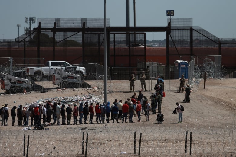 A long line of people stand at a heavily guarded fence and border crossing