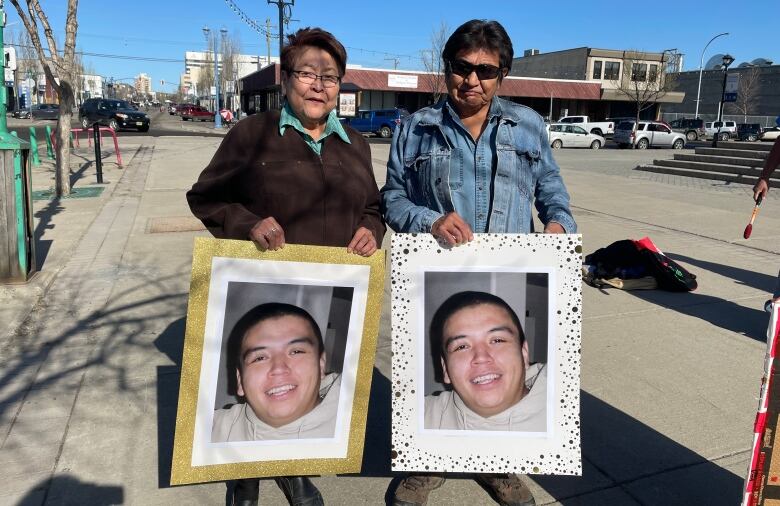 A man and a woman hold portraits of a smiling man outside a building.