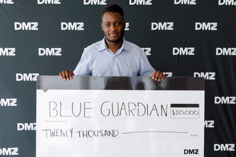 A tall Black man stands in front of banner that says DMZ,  holding up a giant cheque for $20 000.  He won the money for his business. 