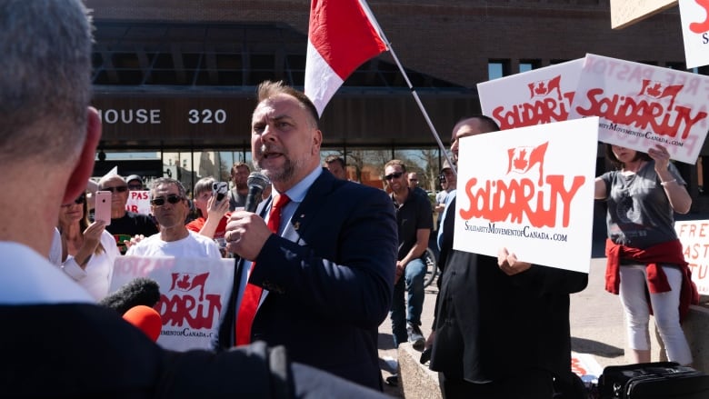 A man in a suit and tie holds a microphone as he speaks to a crowd of supporters and reporters. 