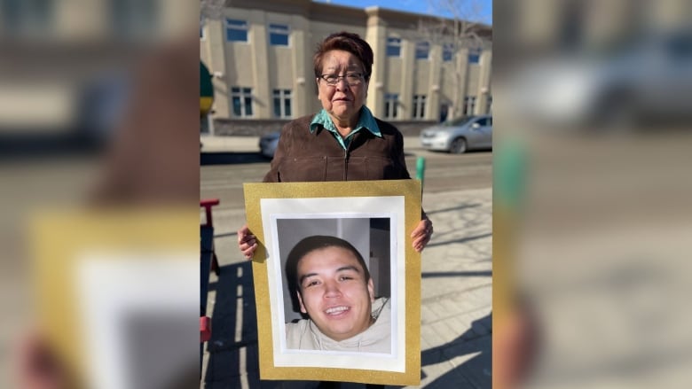 An Indigenous woman holds a framed picture of a smiling man outside a building.