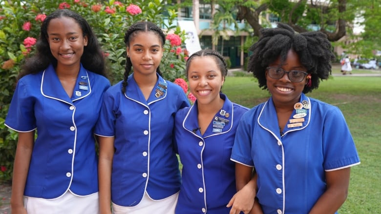 Four Black teen girls, wearing blue and white school uniforms, smile as they pose for a portrait outdoors.