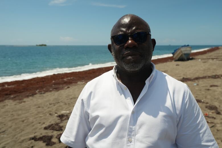 A Black man wearing sunglasses and a white shirt stands on a beach.