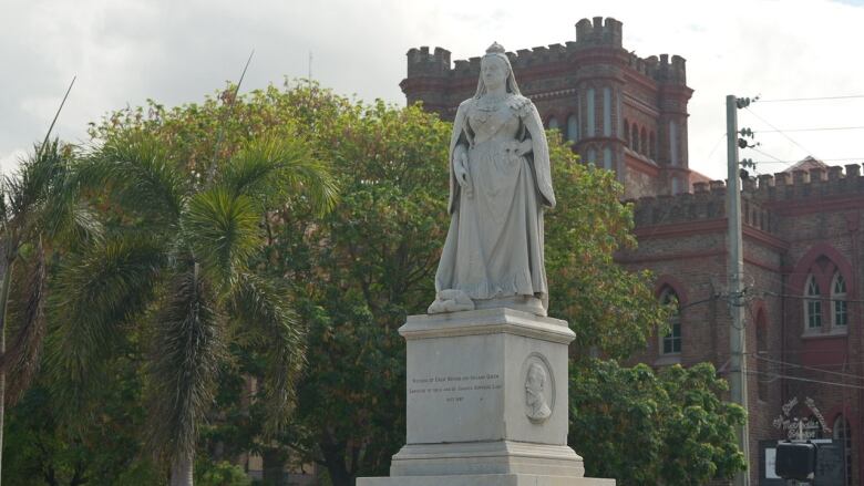 A stone statue of a woman wearing a crown and other regalia is shown, with a red brick building and large tree in the background.