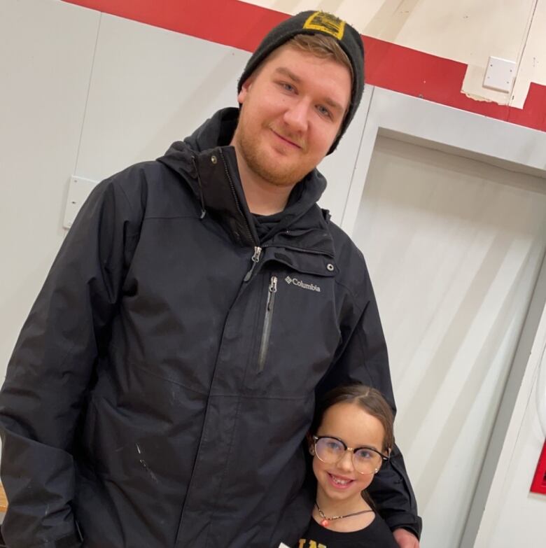 A man in a beanie, black jacket and jeans standing next to a little girl wearing glasses.