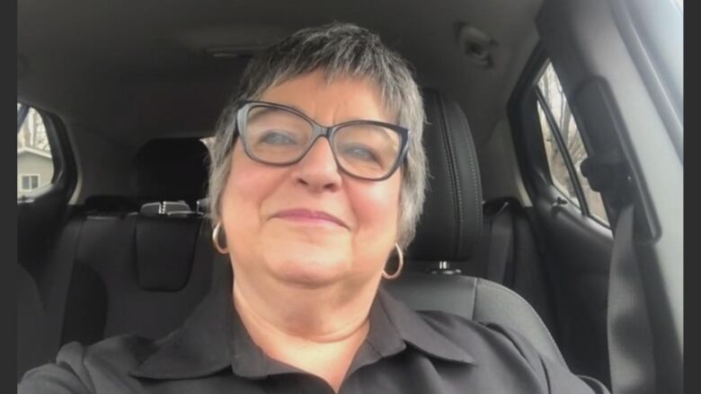 A middle-aged woman with glasses smiles as she sits in her car.