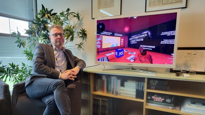 A gray-haired man in black jeans and a blazer sits crossed-legged on an armchair next to a large TV screen displaying the video game Counter-Strike. On the screen, you see a hand holding a gun from a first-person perspective in a room with a large map and the words 