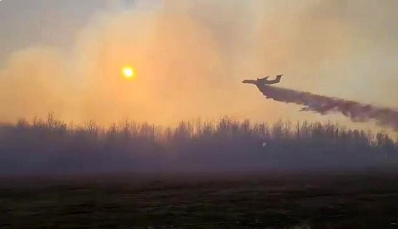 A plane trailing fire retardant flies over treetops with the sun glowing through the rising smoke.