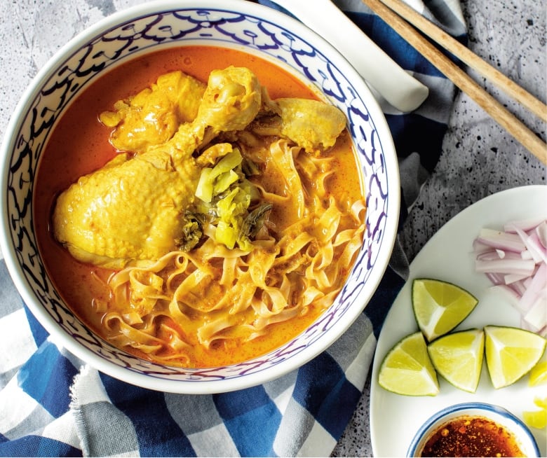 overhead shot of a blue and white bowl filled with a chicken drumstick, noodles and a curry broth. a plate with lime wedges, sliced shallots and a small bowl of chili oil sit next to it. 