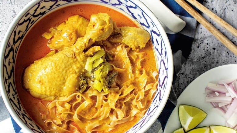 overhead shot of a blue and white bowl filled with a chicken drumstick, noodles and a curry broth. a plate with lime wedges, sliced shallots and a small bowl of chili oil sit next to it. 