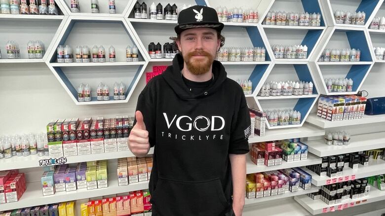 A man with a beard wearing a black hoodie and baseball cap. He is standing in front of a display with different e-cigarette products.