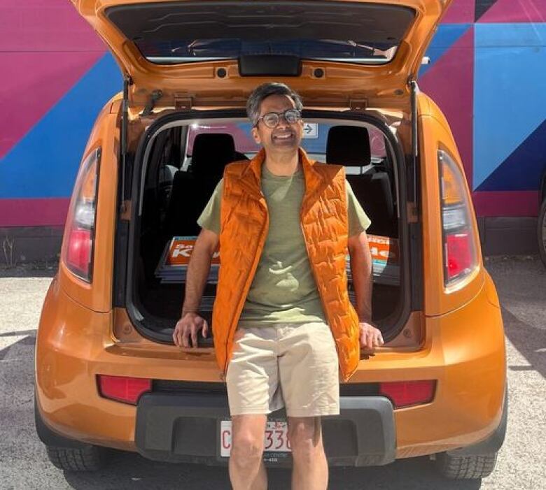 A man leans against the open back of a orange hatchback car. 