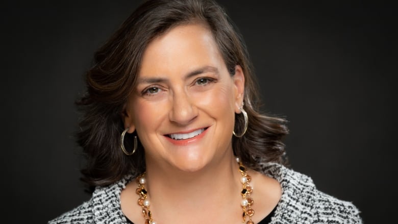 A close up of a woman with brown shoulder-length straight hair looking into the camera and smiling, with a black and white blazer, hoop earrings and a chunky necklace against a black background.