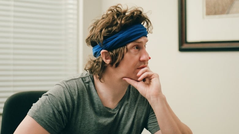 In a still from a movie, a man sits at an office desk, with his head leaning in his hand and an inscrutable expression on his face. He wears a blue headband in his messy hair and a grey T-shirt.