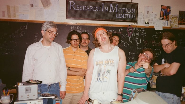 In a still from a movie that takes place in the mid 1990s, 7 young male engineers at Research in Motion stand in a group making silly faces at the camera.