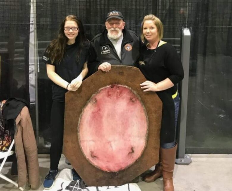 A girl and a woman stand on each side of a man, all three of them holding a beaver pelt.