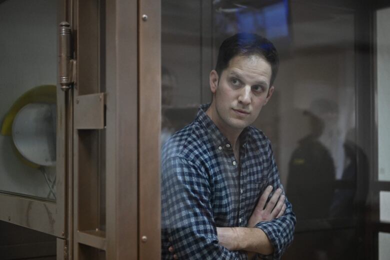 A man wearing a blue plaid shirt stands in a glass box in a courtroom, with his arms crossed in front of his chest.