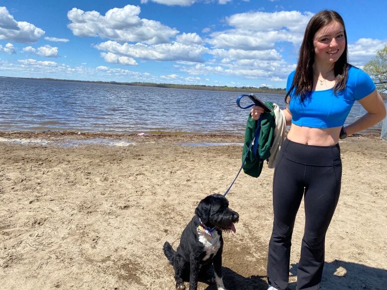 A woman holds a dog leash with the Ottawa River in the background. A black dog with a white belly sits at her heels. 