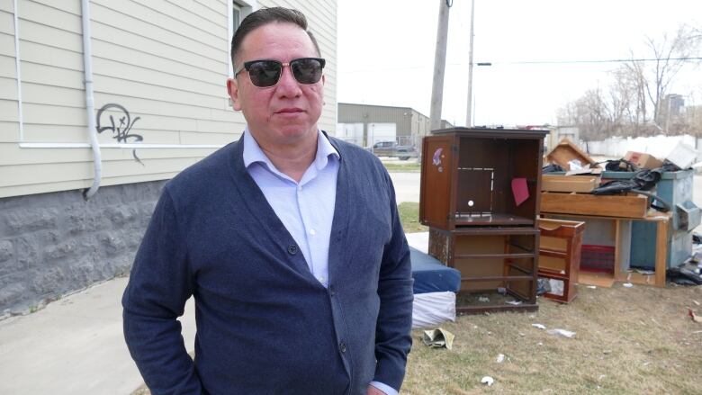 A man in sunglasses and a blue sweater stands in front of a dumpster overflowing with garbage, which is surrounded by various pieces of discarded furniture.