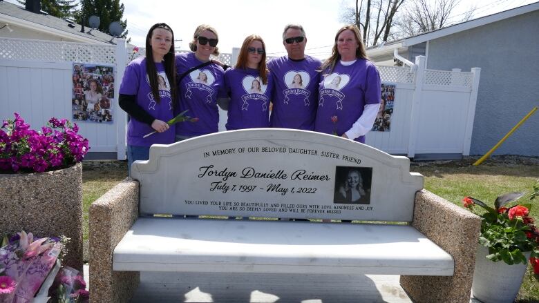 A family is pictured behind a bench engraved with a woman's name, photo and dates of her birth and death.