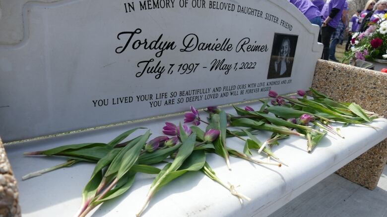 A bench engraved with a woman's photo, name, and dates of birth and death is pictured with flowers placed on it.