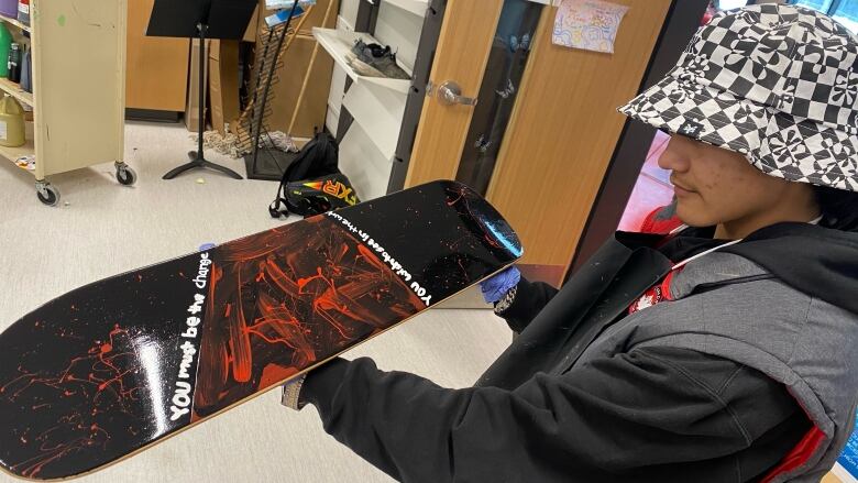 A student holds a painted skateboard. It reads: You be the change you wish to see in the world.