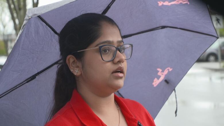 An Indian woman stands under an umbrella and speaks to a camera.