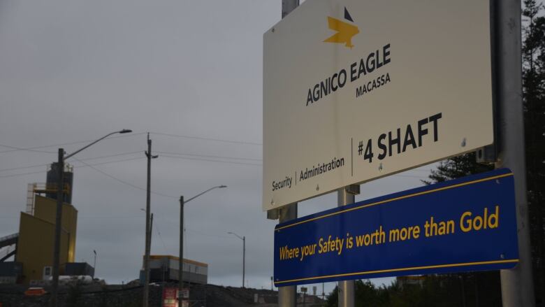 A sign reads 'Agnico Eagle: Macassa' while another reads 'Where your safety is worth more than gold' with mining buildings in the background 