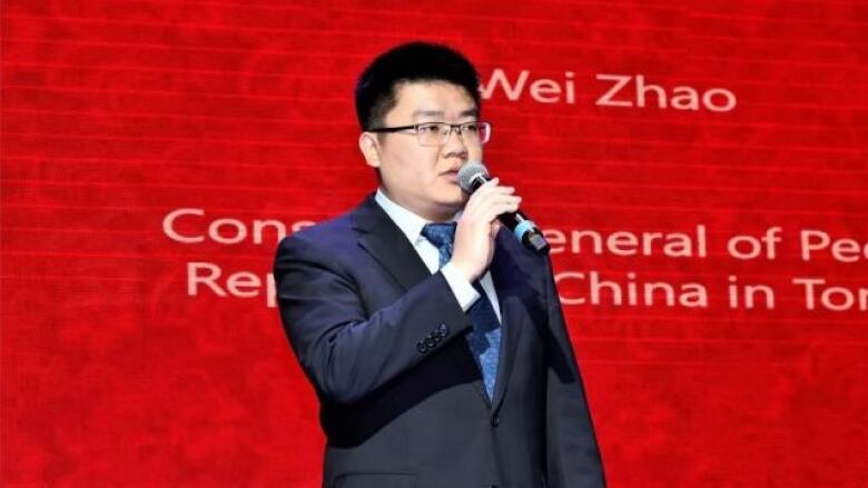 A man in a suit, standing against a red backdrop, speaks into a microphone.