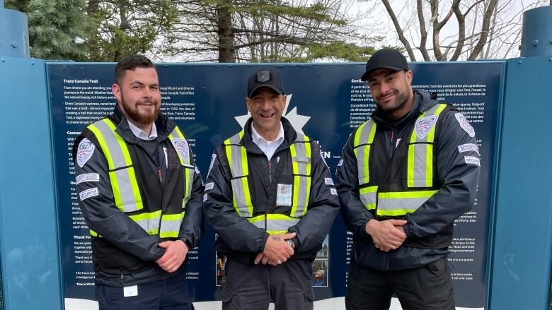 Three uniformed men pose together in front of a trail sign. 