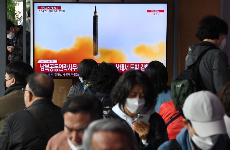 A group of people stand in a queue, with a TV screen showing the firing of a missile in the background.