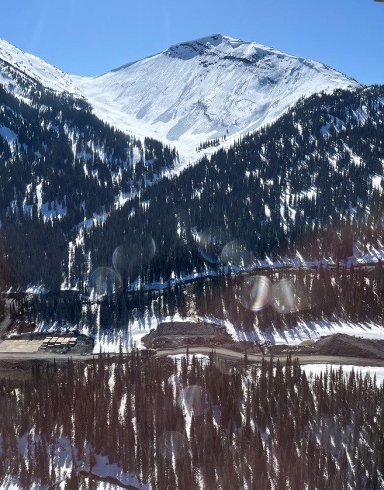 A snow capped mountain in northern B.C. towers over forested hills and the Anzac River, where construction activity is visible along the river banks. 