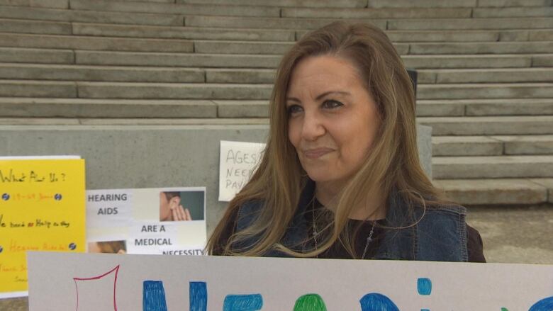 A woman with long brown hair carrying a colourful sign looks off to the side with a slight smile.