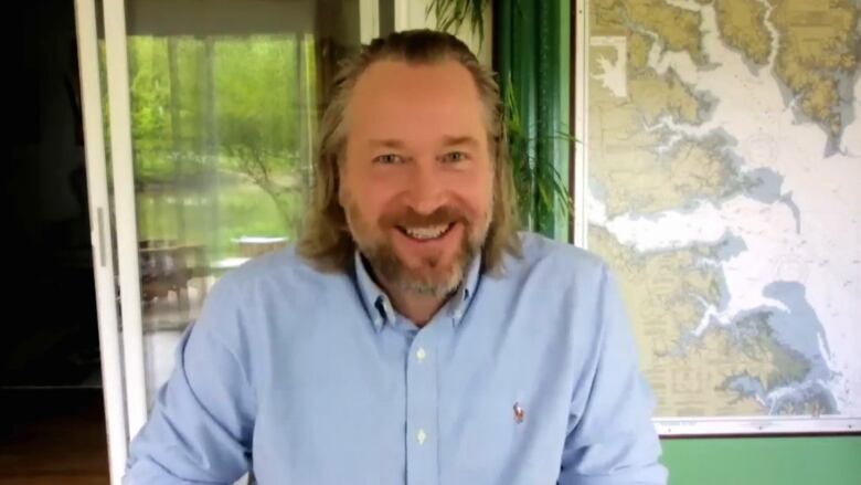 A man with shoulder length hair and a beard smiles in front of a window.