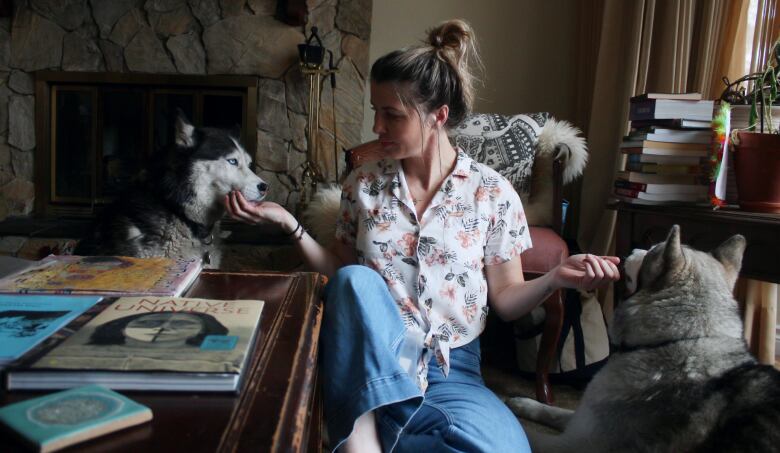 A woman sits on the floor, giving her two huskies treats - on either side of her.