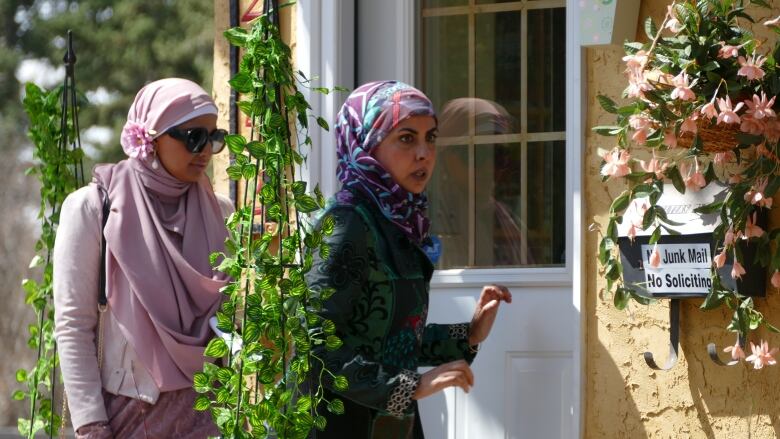 Two women in hijabs stand in front of a yellow house on a spring day. 