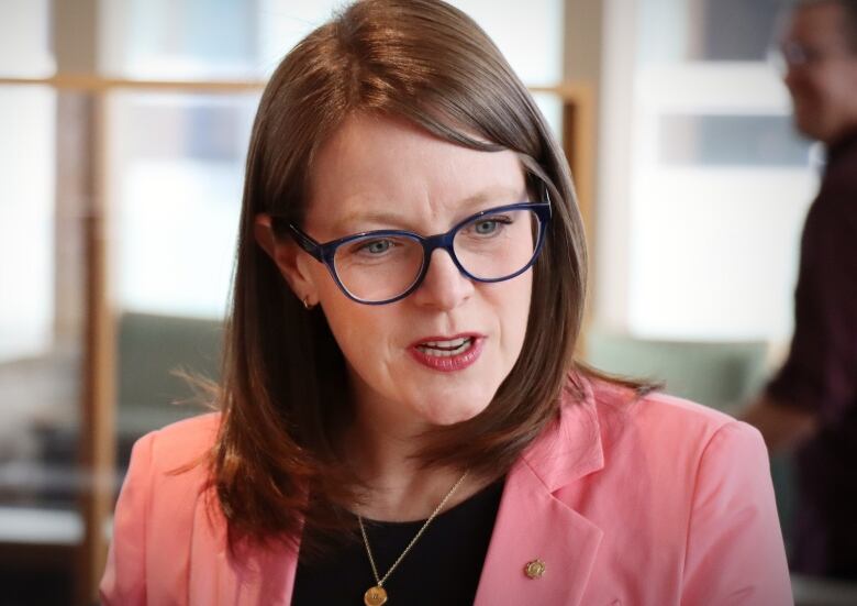 A woman with glasses and a pink suit jacket speaks to someone off camera.