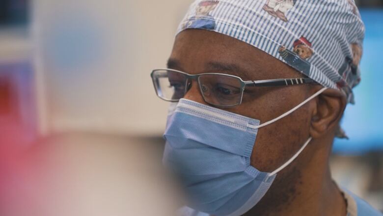 A Black man wearing a blue surgical mask, silver wire-framed glasses and a nurse's cap with a teddy bear print is shown in profile.