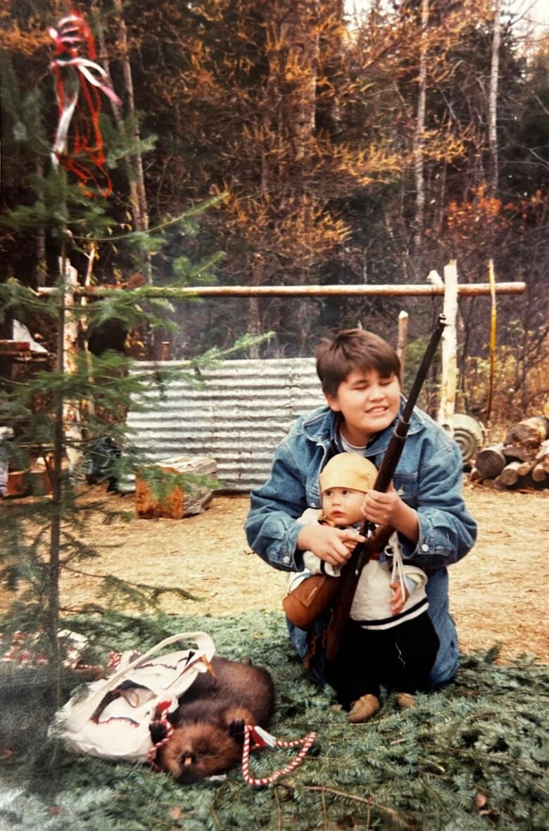 A teenage boy helps a baby dressed in traditional Indigenous clothes hold a rifle on a bed of spruce boughs.