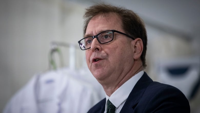 A man wearing glasses speaks in front of lab coats in the background.