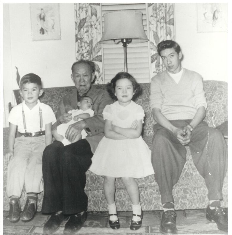 An older man is holding a baby while sitting with three children on a sofa inside an apartment in this black and white photo circa 1955.