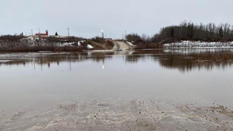 Water floods over a road with snow visible to the sides and a community in the background 