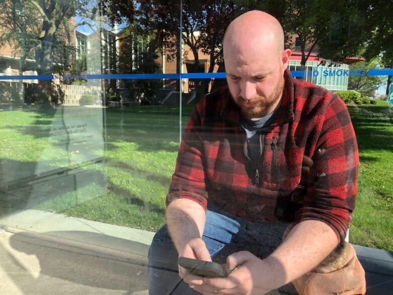 A bald man wearing a red chequered sweater and blue jeans looking at his phone at a bus stop