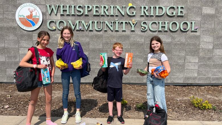 Four students stand outside a school building, holding grocery items like bananas and oranges.