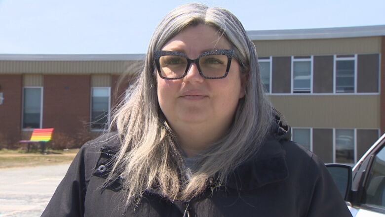 A woman with large glasses stands in front of a school.