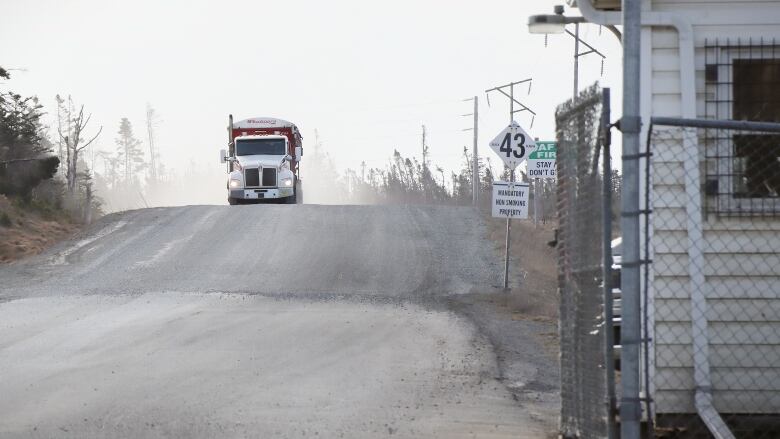 A large semi-truck drives over a small rise on a gravel road heading towards a gate at the entrance to the Donkin mine property.