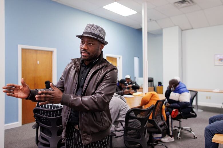 A man in a leather jacket and fedora gestures while speaking. In the background, other people are huddled at a table.