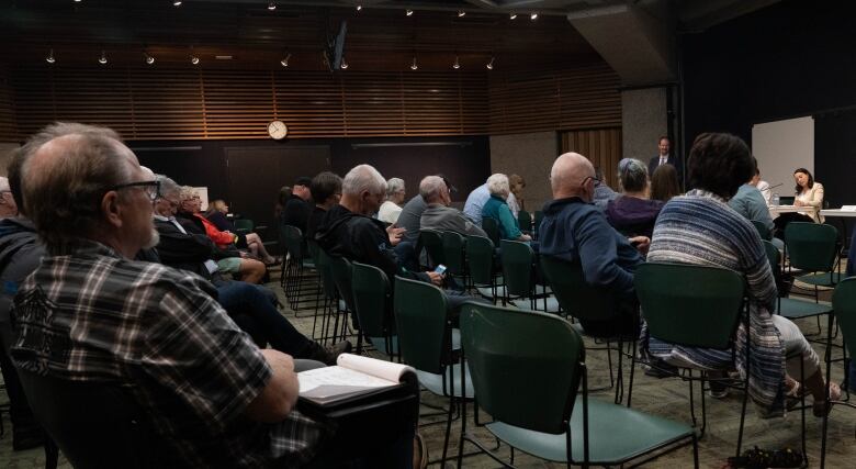 people in green chairs in a room watch speakers sitting in front of a table